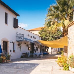 Exterior view of a quaint restaurant in Cyprus with outdoor seating, palm trees, and stone architecture.