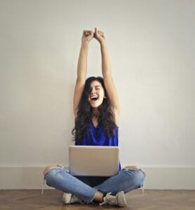 Happy woman with laptop, celebrating success indoors. Arms raised in triumph.