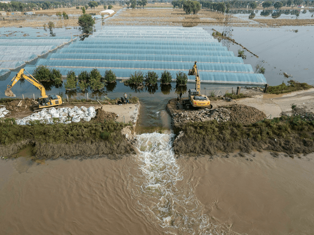 Flooded farmland with submerged greenhouses and excavators managing floodwaters, illustrating climate change impacts on agriculture.