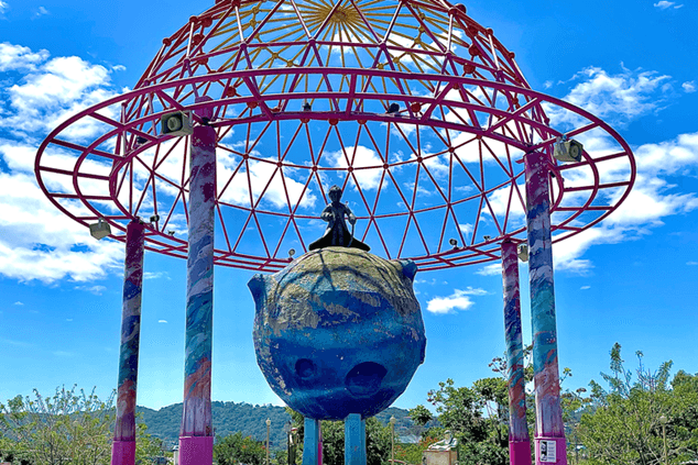 Sculpture of the Little Prince on his small planet under a colorful framework canopy in a themed park setting.