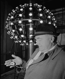 Historical image showing a man holding a pocket watch near the original Times Square New Year's Eve ball with multiple light bulbs, likely related to the first ball drop in 1907.