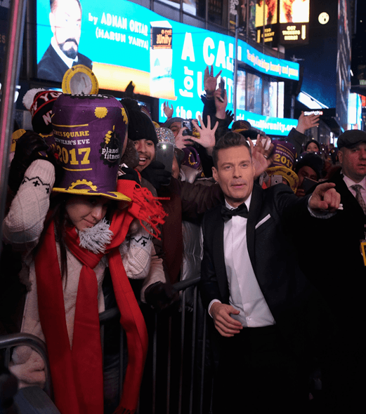 Crowd celebrating New Year's Eve Times Square Ball Drop with festive hats and illuminated billboards in the background.